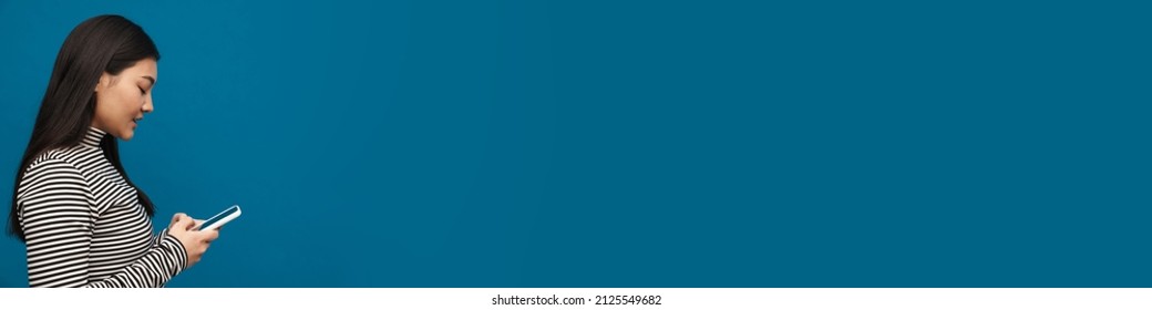 Brunette asian teen girl looking posing and using mobile phone isolated over blue background