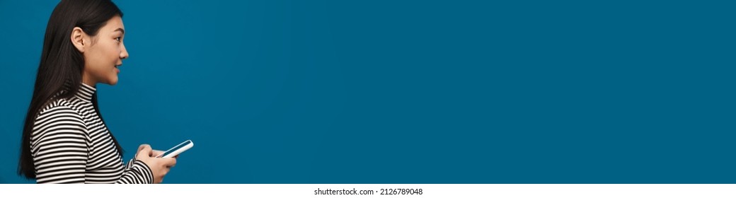 Brunette asian teen girl looking forward while using mobile phone isolated over blue background