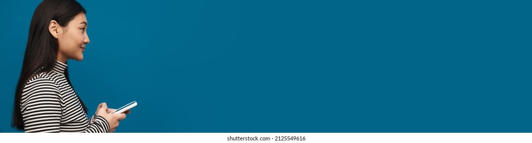 Brunette asian teen girl smiling and using mobile phone isolated over blue background