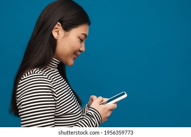 Brunette asian teen girl thinking while using mobile phone isolated over blue background