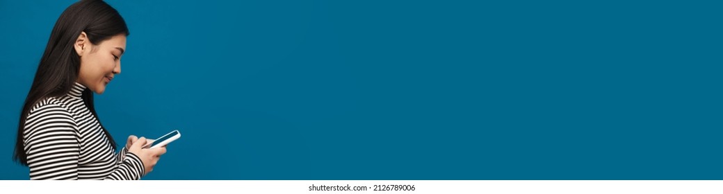 Brunette asian teen girl thinking while using mobile phone isolated over blue background