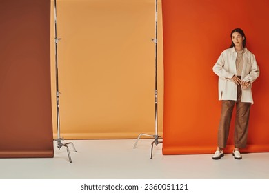 brunette asian woman in autumn attire standing on multicolored backdrop, leather pants and outerwear