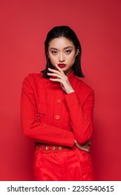 brunette asian woman in fashionable blazer holding hand near face while looking at camera isolated on red - Shutterstock ID 2235540615