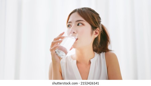 Brunette Asian Woman With Brunette Ponytail Drink A Glass Of Water In A White Room