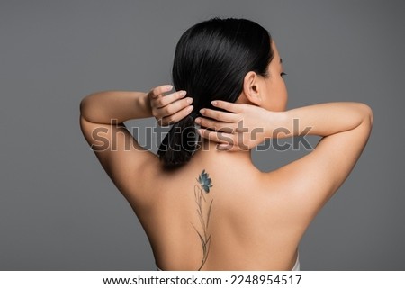 Brunette asian woman with tattoo on back touching ponytail isolated on grey