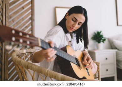 Brunette Attractive Female Learning To Play Guitar While Sitting In Cozy Home Interior. Young Concentrated Woman Enjoying Her Hobby With Musical Instrument Performance Relaxing On Wicker Chair