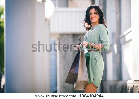 brunette with a beautiful figure and smile is standing in summer tunic with packages and eyeglasses in her hands at day in the street