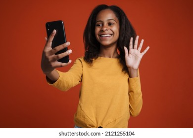 Brunette Black Girl Waving Hand And Using Mobile Phone Isolated Over Red Wall