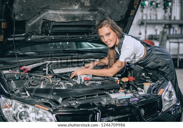 A brunette in a black jumpsuit and a\
white t-shirt near the open hood of black car. Young female in the\
garage is smiling at the camera. car repair\
concept