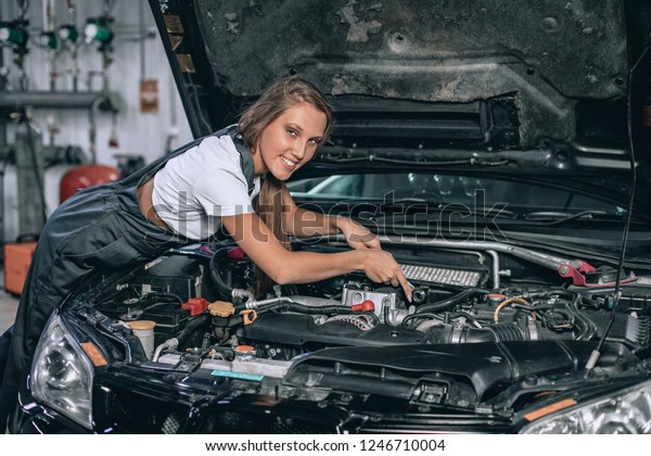 A brunette in a black jumpsuit and a
white t-shirt near the open hood of black car. Young female in the
garage is smiling at the camera. car repair
concept