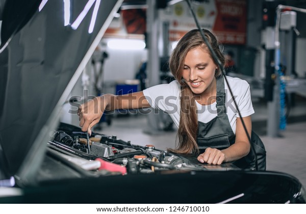 A brunette in a\
black jumpsuit and a white t-shirt near the open hood of black car.\
Young female in the garage is smiling at the camera and lowered\
gaze. car repair concept