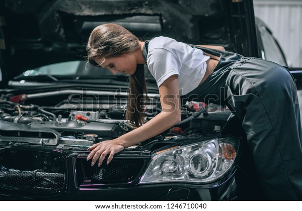 A brunette in a\
black jumpsuit and a white t-shirt near the open hood of black car.\
Young female in the garage is smiling at the camera and lowered\
gaze. car repair concept