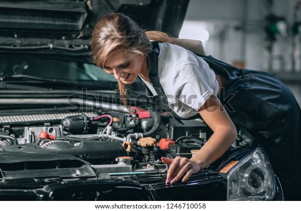 A brunette in a
black jumpsuit and a white t-shirt near the open hood of black car.
Young female in the garage is smiling at the camera and lowered
gaze. car repair concept