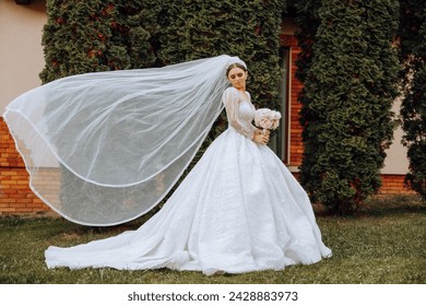 A brunette bride in a voluminous white dress holds a bouquet, poses. The veil is thrown into the air. Beautiful hair and makeup. Wedding photo session in nature. Long train of the dress - Powered by Shutterstock