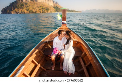 brunette bride in white wedding dress and handsome groom kiss in longtail boat drifting  to island at sunrise