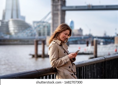 A Brunette Buisness Woman In A Trench Coat Reads Emails On Her Mobile Phone In Front Of The Skyline Of London, United Kingdom