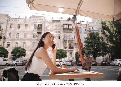 Brunette Business Asian Girl Sits At Table In Cafe Against Backdrop Of City Building Under Umbrella. She Is On Phone And Looks Away With Smile. On Table Is Laptop, Glass Of Coffee. Freelancer Concept