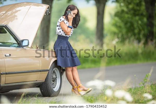 Brunette in\
business clothes and bonnet open leans against the broken down car,\
calling for help and\
assistance.