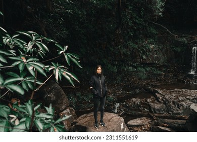 brunette caucasian girl in black jacket dark trousers and sneakers smiling at camera standing on big rock among waterfalls and forest vegetation,purakaunui falls, new zealand - Shutterstock ID 2311551619