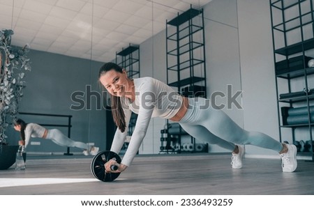 Brunette cheerful young woman in sportswear at abdominal exercise, rollouts. Fit caucasian girl improving body endurance, Happy fitness model at workout, looks air camera toothy smiles.