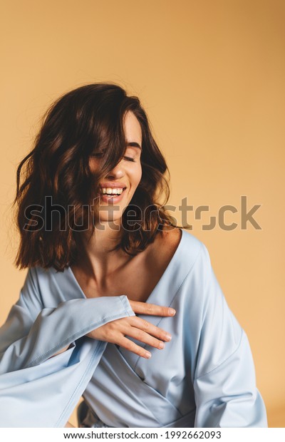 Brunette with\
curls on a beige background, natural make-up and clean skin.\
Beautiful smile and closed eyes. Portrait laughing sensual woman.\
Girl wear fresh blue color kimono\
dress.