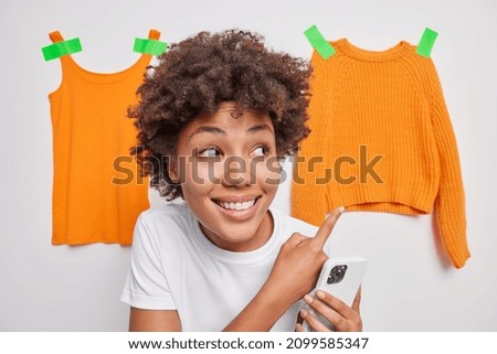Brunette curly haired woman points at clothes hanging on white background suggests discount holds mobile phone sells unnecessary items of clothing online. Look at this orange knitted sweater