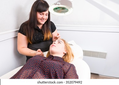 Brunette Esthetician Cleaning Up Excess Dye Coloring From Eyebrows Of Blond Female Client Reclined On Chair In White Spa