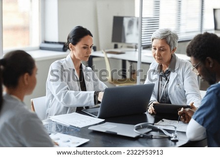 Brunette female clinician in lab coat pointing at laptop screen while making presentation of medical data to group of intercultural colleagues