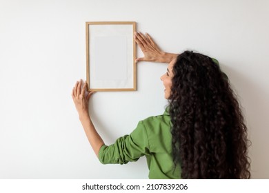Brunette Female Hanging Empty Picture In Frame On White Wall Decorating House Standing Indoors. Modern Art And Home Interior Decor. Selective Focus On Blank Poster. Mockup