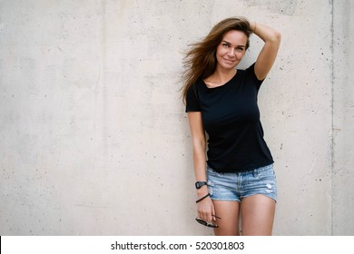 A brunette female with long hair wearing a blank black t-shirt is smiling at the camera while standing on a concrete wall background on a street. Empty space for text or design.