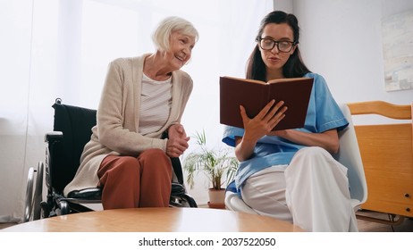 Brunette Geriatrician Nurse Reading Book To Happy Aged Woman In Wheelchair