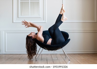 A Brunette Girl In Black Leggings Lies On A Black Chair And Does Yoga Exercises. Background White