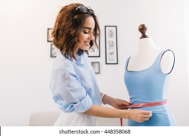 A brunette girl in a blue shirt is working in the workshop studio. She makes fitting on the blue dress on the  mannequin.