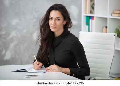 A brunette girl in a business black dress is sitting at a table in the office and is working hard. Student writes notes, takes notes in a notebook