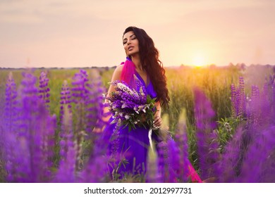 A brunette girl in gradient haute couture dress in blue  pink   yellow colors standing among blooming lupine field  A magical romantic sunset portrait 