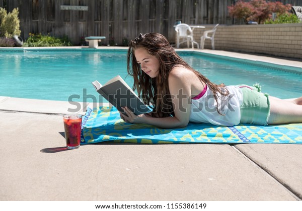 Brunette girl laying on her stomach in shorts and tank top reading a novel ...