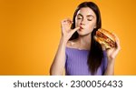 Brunette girl licks her finger, eats tasty hamburger. Woman orders takeaway food with delivery app, enjoys delicious burger, orange background. Restaurants and takeout concept