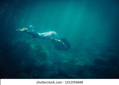 brunette girl in long blue dress dives underwater in the sea