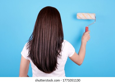 Brunette Girl With Long Hair Using Paint Roller, View From Behind. Isolated On Blue Background.