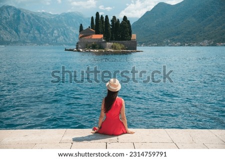 Brunette girl in red dress and hat sitting in front of scenic lake or bay in Perast, Montenegro, a top Europe travel destination. Summer travel guide vacation background with people from behind