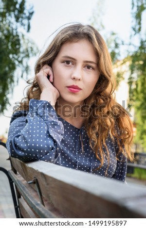 Brunette girl sitting on bench in the city. Portrait of girl in blue dress with long hair on the street in summer day