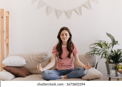 Brunette girl in striped T-shirt is meditating while sitting on sofa in living room. Calm woman in red tee relaxing on beige couch at home - Powered by Shutterstock