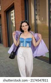 Brunette Girl Walks In The City. Light Trousers, Blue Corsage, Lilac Jacket