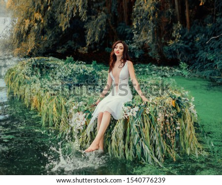 brunette girl in a white vintage dress with a deep neckline, sitting on a boat and playing bare feet in river water. Spray and drops. Cute face. Summer day. Wedding decorations with willow and flowers