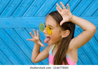A brunette girl in yellow sunglasses stands against a blue wall of planks and makes a funny grimace, showing her tongue. Naughty ill-mannered child