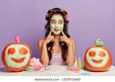 Brunette Glad Asian Woman Applies Facial Mask, Points Both Index Fingers On Face, Wears Curlers, Has Beauty Procedures, Two Funny Ripe Pumpkins With Drawn Faces On White Desk Near. Halloween Is Soon