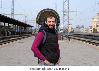 Brunette guy at an empty train station. The train station without people. The guy in the jacket and black sleeveless jacket is looking at the camera. Waiting for a train to travel. 