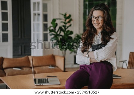 Brunette hispanic businesswoman in glasses sitting on desk with laptop smiles wide looks aside at break of work at office. Successful people. Happy accountant remote working home.