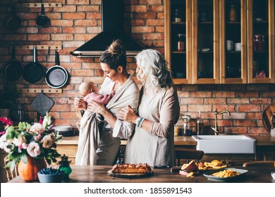 Brunette With Holds Baby And Senior Lady Looking To The Granddaughter Preparing Dinner In Kitchen. 