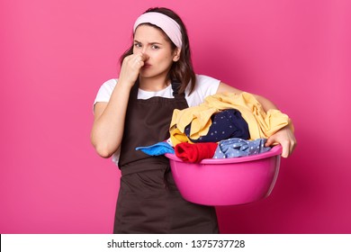 Brunette Houseworker Holding Her Nose With One Hand, Having Full Pink Basin Of Bad Smelling Items Of Clothes, Trying To Bear Smell. Emotional Exhausted Model Poses Isolated Over Pink Background.